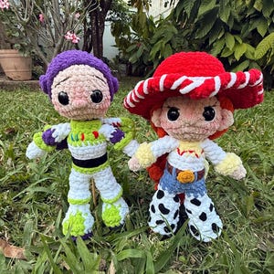 May include: Two handmade crochet dolls of cartoon characters, Buzz Lightyear and Jessie. Buzz is in a green, white, and purple spacesuit. Jessie wears a red hat with white trim, a white shirt, blue jeans, and cow-print trousers. They are on green grass.