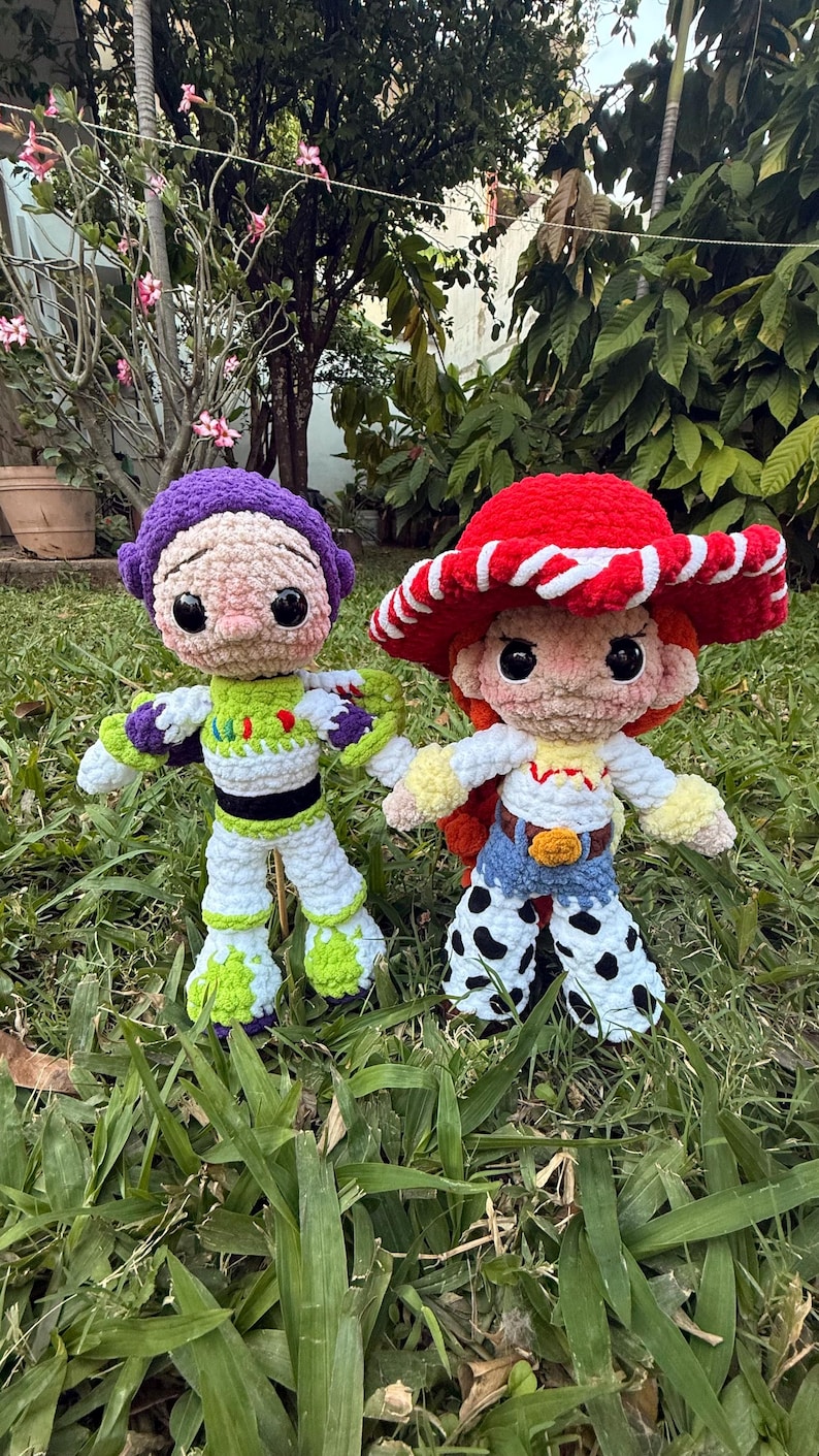 May include: Two handmade crochet dolls of cartoon characters, Buzz Lightyear and Jessie. Buzz is in a green, white, and purple spacesuit. Jessie wears a red hat with white trim, a white shirt, blue jeans, and cow-print trousers. They are on green grass.