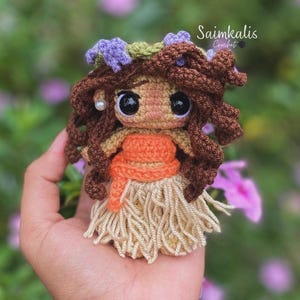 K&ouml;nnte beinhalten: Eine handgefertigte H&auml;kelpuppe mit braunen Locken, gro&szlig;en schwarzen Augen und einem beigen Rock. Die Puppe hat ein orangefarbenes Oberteil und einen Blumenkranz. Die Puppe wird in einer Hand gehalten, mit dem Text "Saimkalis Crochet" im Hintergrund.