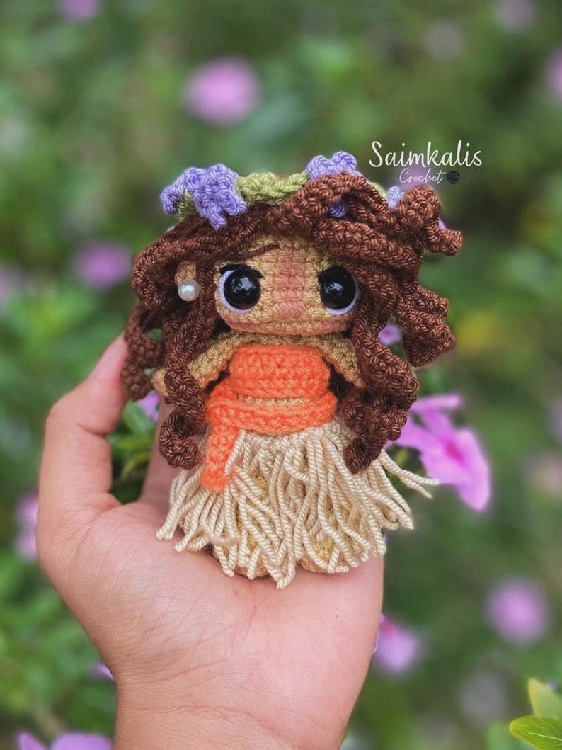 K&ouml;nnte beinhalten: Eine handgefertigte H&auml;kelpuppe mit braunen Locken, gro&szlig;en schwarzen Augen und einem beigen Rock. Die Puppe hat ein orangefarbenes Oberteil und einen Blumenkranz. Die Puppe wird in einer Hand gehalten, mit dem Text "Saimkalis Crochet" im Hintergrund.