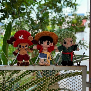 Op de afbeelding: Drie handgemaakte gehaakte figuren van personages uit de One Piece anime-serie. De figuren staan op een metalen hek. De personages zijn Tony Tony Chopper, Monkey D. Luffy en Roronoa Zoro, elk met verschillende outfits en kenmerken.