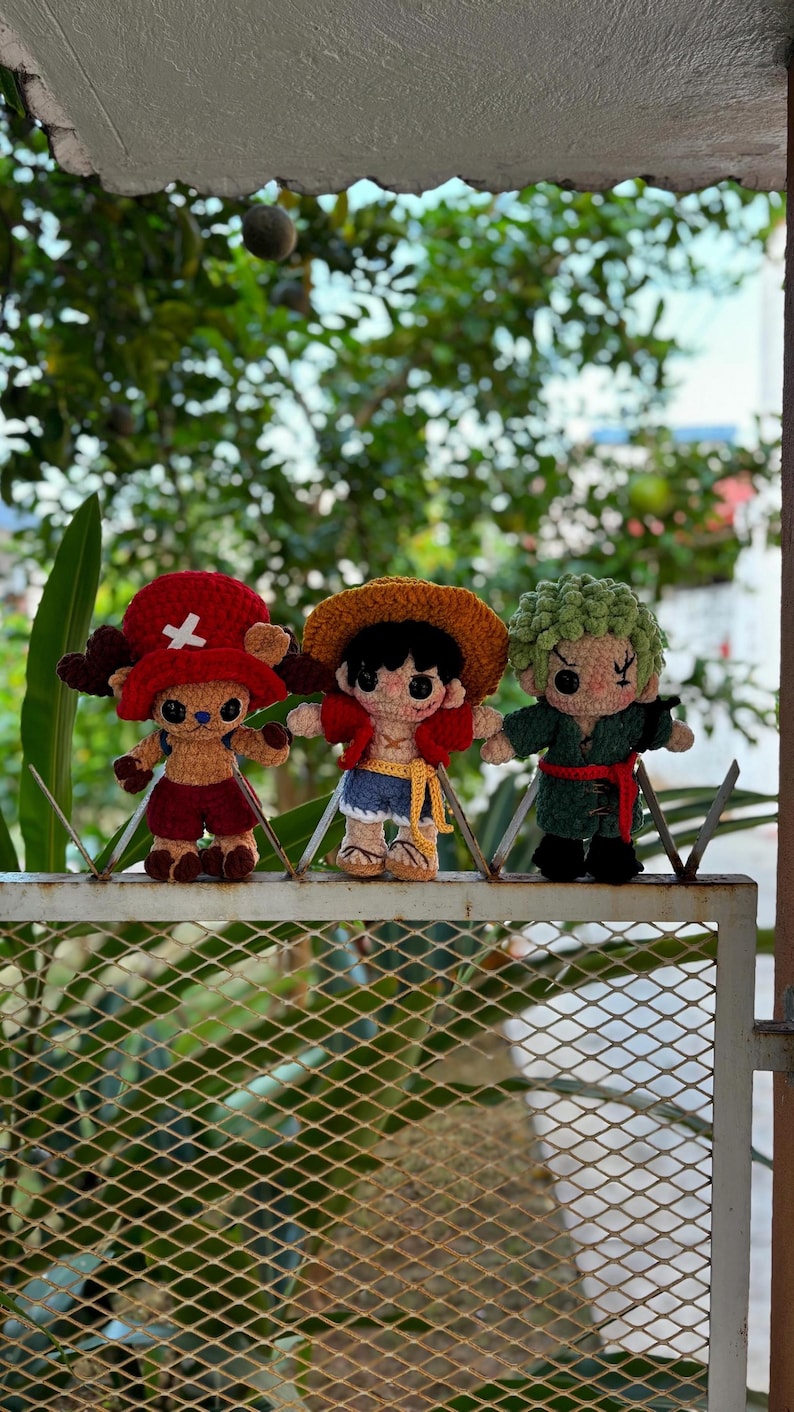 Op de afbeelding: Drie handgemaakte gehaakte figuren van personages uit de One Piece anime-serie. De figuren staan op een metalen hek. De personages zijn Tony Tony Chopper, Monkey D. Luffy en Roronoa Zoro, elk met verschillende outfits en kenmerken.