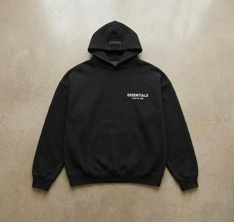 May include: Black hooded sweatshirt with a front pocket and the word "ESSENTIALS" printed in white. The sweatshirt has a drawstring hood and a small label on the hood.