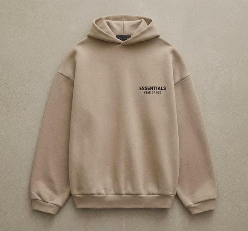 May include: A tan hoodie with a drawstring hood and long sleeves. The text "ESSENTIALS FEAR OF GOD" is printed on the upper left chest. The hoodie is made of a soft, comfortable material.