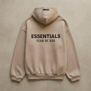 May include: A tan hoodie with the text "ESSENTIALS FEAR OF GOD" printed in black on the back. The hoodie has a hood and long sleeves. The background is a solid, neutral color.