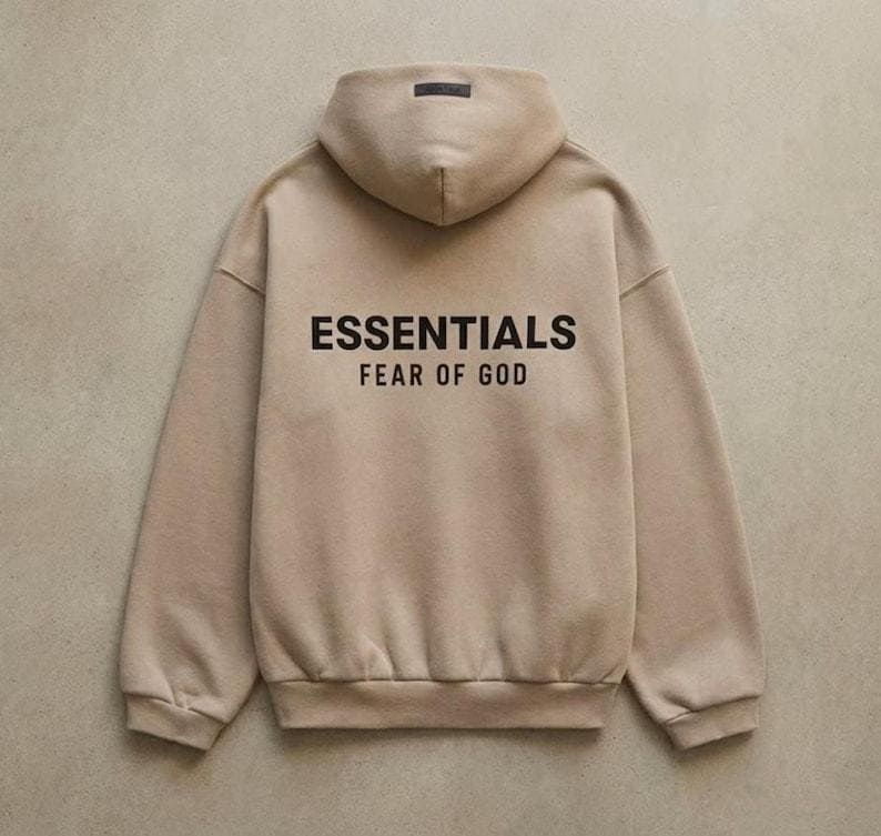 May include: A tan hoodie with the text "ESSENTIALS FEAR OF GOD" printed in black on the back. The hoodie has a hood and long sleeves. The background is a solid, neutral color.