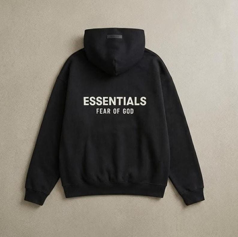 May include: Black hoodie with the word "ESSENTIALS" in white block letters across the back, with "FEAR OF GOD" below it. The hoodie has a hood and long sleeves. The background is a neutral beige color.