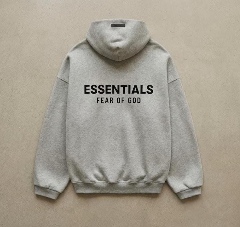 May include: A gray hoodie with the text "ESSENTIALS FEAR OF GOD" printed in black. The hoodie has a hood and a small black label at the top of the hood.