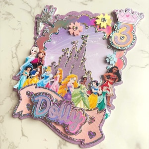 May include: A decorative cake topper featuring Disney princesses, a purple castle, and the number 3. The design includes the name "Dolly" in a banner, with glitter and floral embellishments. The colour scheme is predominantly purple and pink.