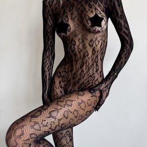 May include: Black lace bodysuit with a high neckline and long sleeves. The bodysuit features a snakeskin pattern and is worn with black star-shaped pasties. The garment is form-fitting and covers the entire body.