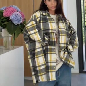 Puede incluir: Una persona lleva una camisa de franela a cuadros amarilla, gris y blanca sobre una camiseta blanca y unos vaqueros azules. La camisa tiene un diseño clásico con botones. Un ramo de hortensias rosas y azules está en el fondo.