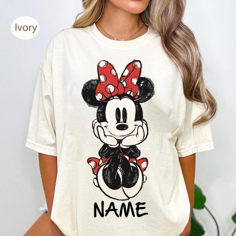 May include: Ivory-colored t-shirt featuring a graphic of Minnie Mouse. Minnie is depicted in black and white with a red polka-dot bow. The word "NAME" is printed in black at the bottom of the graphic.
