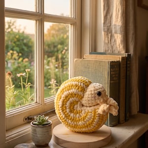 May include: A crocheted amigurumi ammonite plush toy featuring a spiral shell pattern in alternating yellow and cream yarn. The creature has a soft, rounded head with a single visible black eye and small, textured tentacles extending from the front.