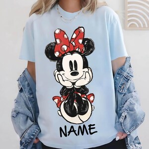 May include: Light blue t-shirt featuring a Minnie Mouse graphic. The graphic shows Minnie Mouse with a red polka dot bow, black ears, and a red polka dot skirt. The word "NAME" is printed in black at the bottom of the shirt.