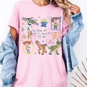 May include: Pink t-shirt featuring cartoon characters from the Toy Story franchise. The design includes Buzz Lightyear, Woody, an alien, and other characters, with the text "YOU'VE GOT A FRIEND IN ME".