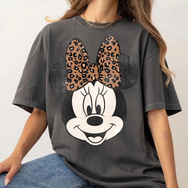 May include: Dark gray t-shirt featuring a Minnie Mouse graphic. The graphic includes a black and white Minnie Mouse face with a leopard print bow. The shirt has short sleeves and a relaxed fit, suitable for casual wear.