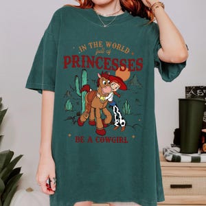 May include: Dark teal t-shirt with a graphic of a cartoon cowgirl hugging a horse, with desert scenery. Text reads "In the world full of princesses, be a cowgirl."