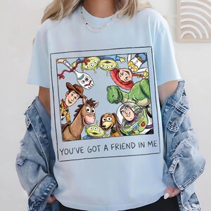 K&ouml;nnte beinhalten: Hellblaues T-Shirt mit einem gerahmten Aufdruck von Toy Story-Charakteren, darunter Woody, Buzz Lightyear und Rex. Der Text "YOU'VE GOT A FRIEND IN ME" ist darunter gedruckt. Das Shirt hat kurze &Auml;rmel und einen Rundhalsausschnitt.