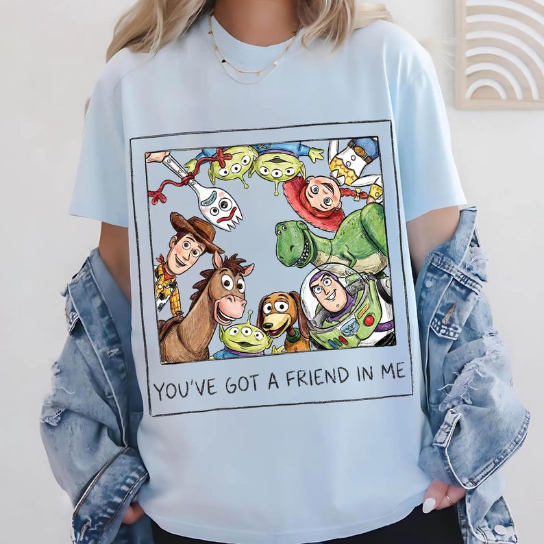 K&ouml;nnte beinhalten: Hellblaues T-Shirt mit einem gerahmten Aufdruck von Toy Story-Charakteren, darunter Woody, Buzz Lightyear und Rex. Der Text "YOU'VE GOT A FRIEND IN ME" ist darunter gedruckt. Das Shirt hat kurze &Auml;rmel und einen Rundhalsausschnitt.