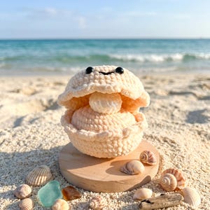 May include: A cream-colored crocheted clam shell with a small round pearl inside, featuring two black bead eyes and a stitched smile. The clam is resting on a circular wooden base, surrounded by small seashells and pieces of sea glass.