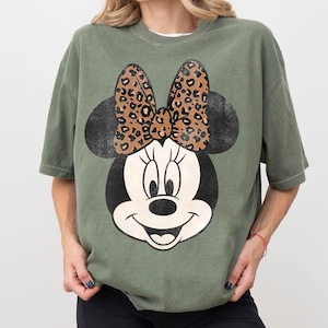 May include: An olive green t-shirt featuring a large graphic of Minnie Mouse. The Minnie Mouse design includes a black head and ears, a white face, and a bow with a leopard print pattern.