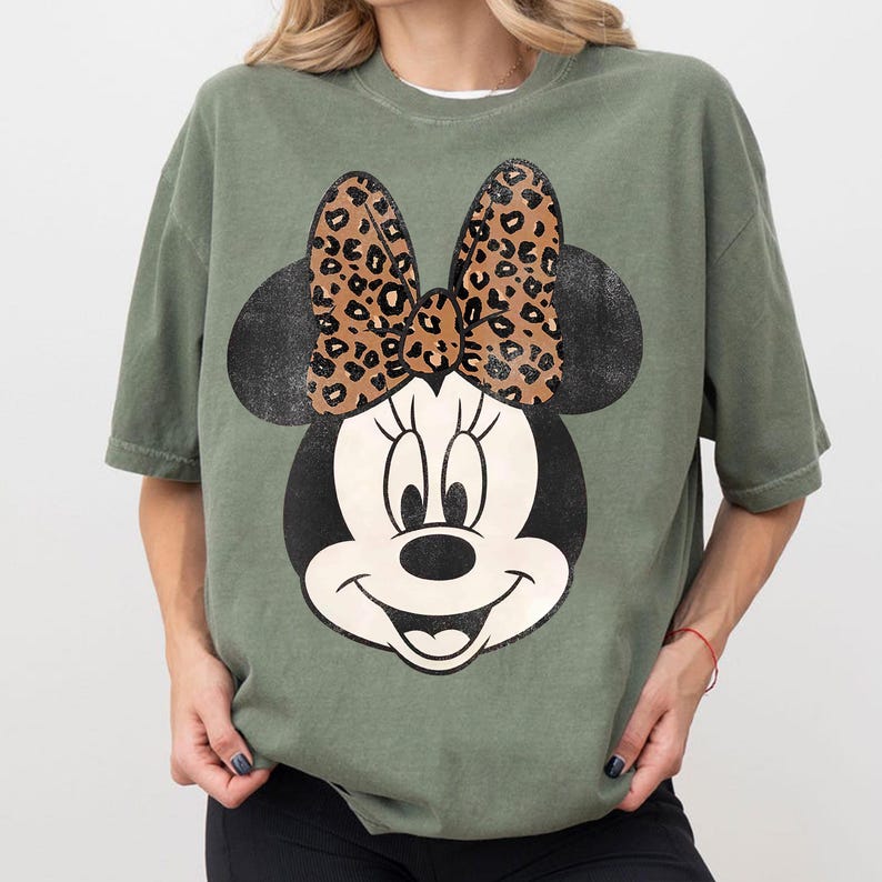May include: An olive green t-shirt featuring a large graphic of Minnie Mouse. The Minnie Mouse design includes a black head and ears, a white face, and a bow with a leopard print pattern.