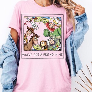 K&ouml;nnte beinhalten: Hellrosa T-Shirt mit einer gerahmten Grafik von Toy Story-Charakteren, darunter Woody, Buzz Lightyear und Rex. Der Text "YOU'VE GOT A FRIEND IN ME" ist darunter gedruckt. Das T-Shirt ist aus weichem Material.