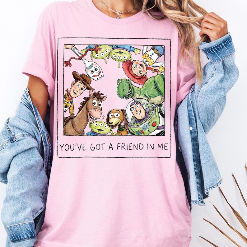 K&ouml;nnte beinhalten: Hellrosa T-Shirt mit einer gerahmten Grafik von Toy Story-Charakteren, darunter Woody, Buzz Lightyear und Rex. Der Text "YOU'VE GOT A FRIEND IN ME" ist darunter gedruckt. Das T-Shirt ist aus weichem Material.
