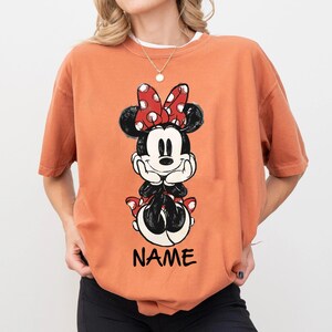 May include: Orange t-shirt featuring a Minnie Mouse graphic. The graphic shows Minnie Mouse with a red polka dot bow, black ears, and the word "NAME" in black block letters. The shirt has short sleeves and a crew neck.