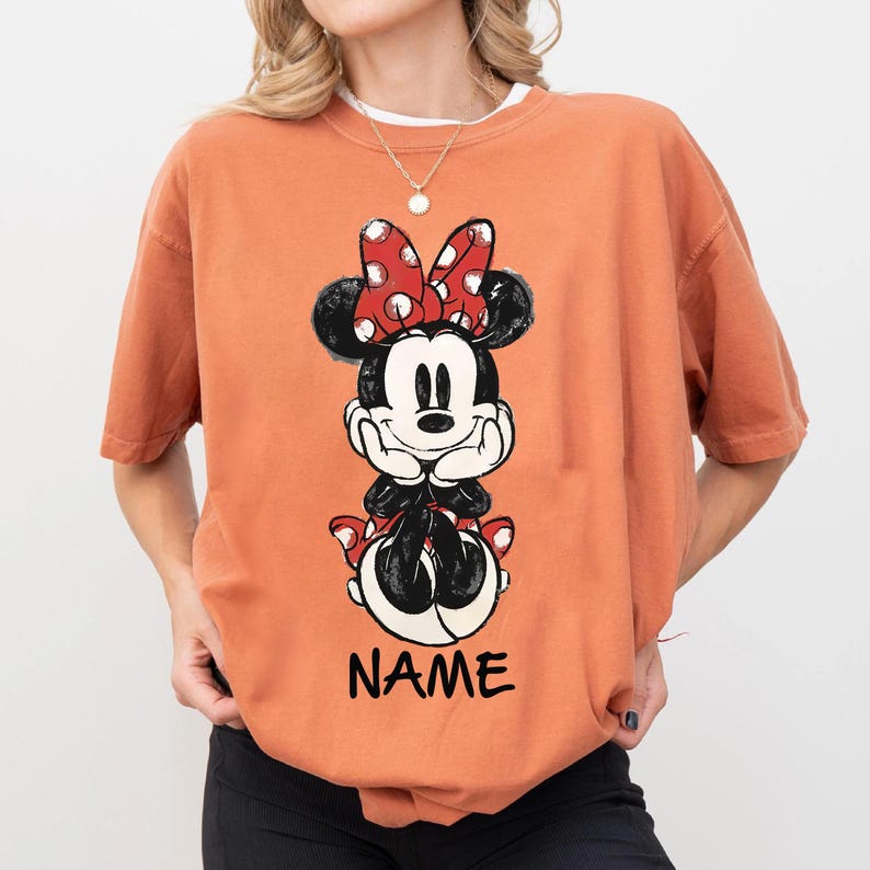 May include: Orange t-shirt featuring a Minnie Mouse graphic. The graphic shows Minnie Mouse with a red polka dot bow, black ears, and the word "NAME" in black block letters. The shirt has short sleeves and a crew neck.