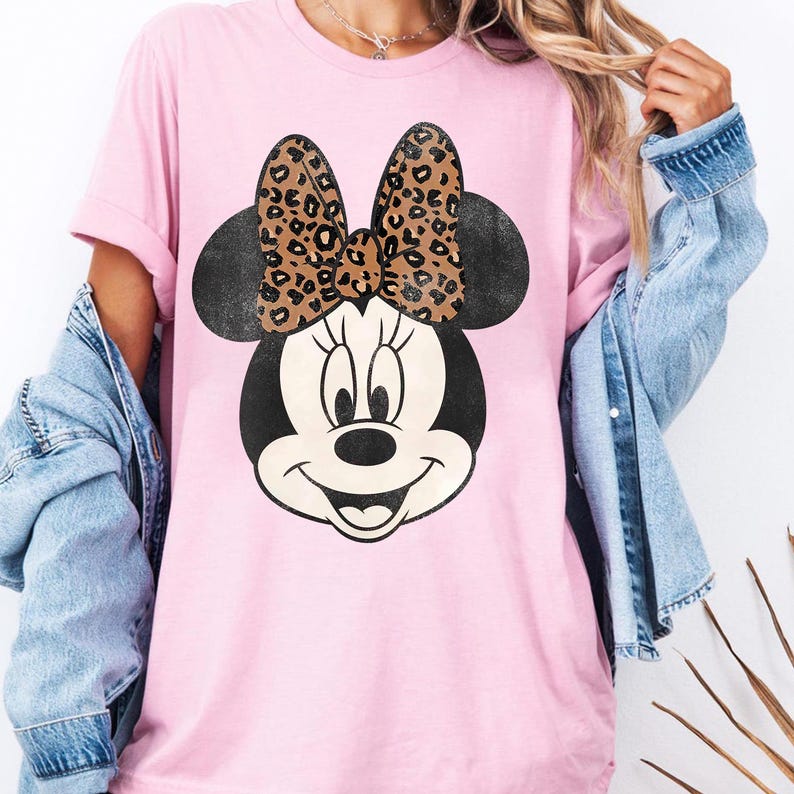 May include: Pink t-shirt featuring a Minnie Mouse graphic. The graphic shows Minnie's face with a black head and ears, a smiling expression, and a bow with a leopard print pattern. The shirt has short sleeves.