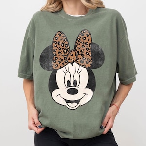 May include: Olive green t-shirt featuring a distressed Minnie Mouse graphic. The graphic includes Minnie's face, black ears, and a leopard print bow. The shirt has a relaxed fit and short sleeves.