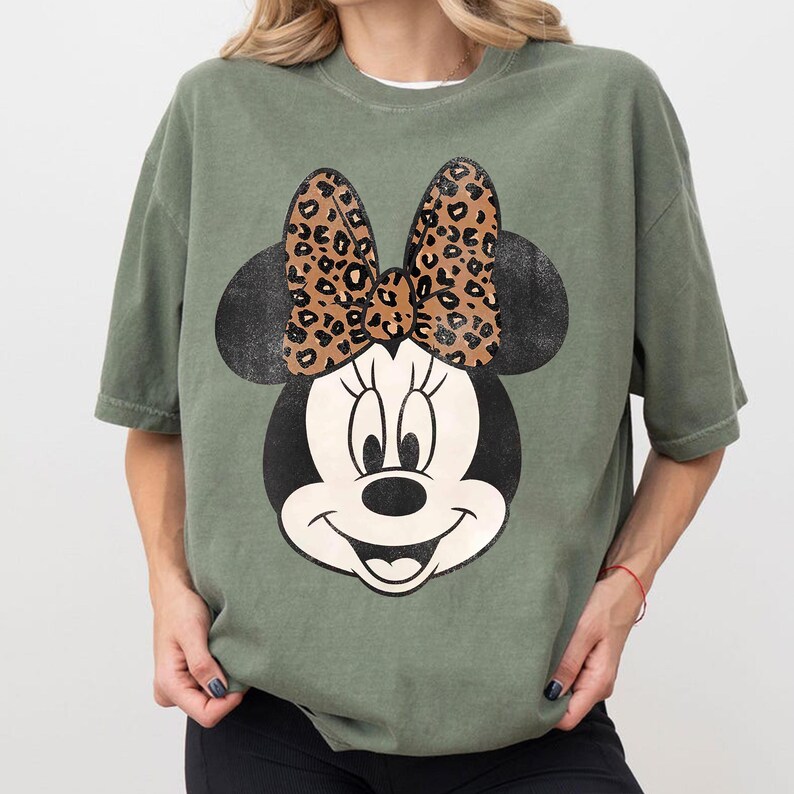 May include: Olive green t-shirt featuring a distressed Minnie Mouse graphic. The graphic includes Minnie's face, black ears, and a leopard print bow. The shirt has a relaxed fit and short sleeves.