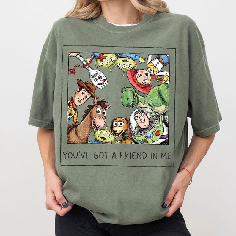 K&ouml;nnte beinhalten: Olivgr&uuml;nes T-Shirt mit einer gerahmten Illustration von Toy Story-Charakteren, darunter Woody und Buzz Lightyear. Die Illustration ist von einem schwarzen Rahmen umgeben. Der Text "YOU'VE GOT A FRIEND IN ME" steht darunter.