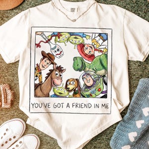 May include: Cream-colored t-shirt featuring a colorful illustration of characters from the movie "Toy Story." The graphic includes Woody, Buzz Lightyear, Rex, and other characters, with the text "YOU'VE GOT A FRIEND IN ME" below the image. The shirt is paired with white sneakers.