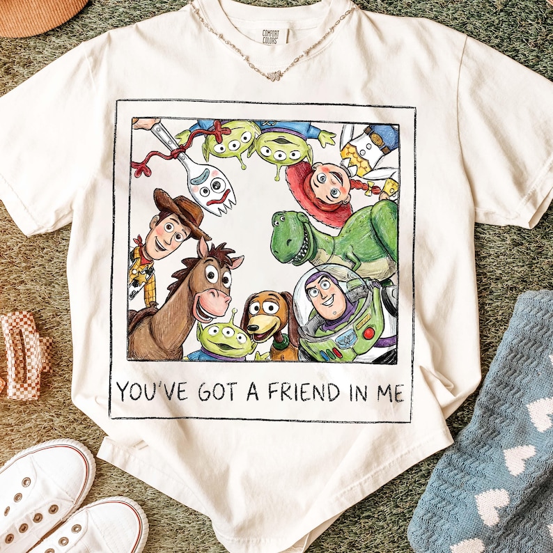 K&ouml;nnte beinhalten: Cremefarbenes T-Shirt mit einer farbenfrohen Illustration von Charakteren aus dem Film "Toy Story". Die Grafik zeigt Woody, Buzz Lightyear, Rex und andere Charaktere mit dem Text "YOU'VE GOT A FRIEND IN ME" darunter. Das Shirt wird mit wei&szlig;en Turnschuhen kombiniert.