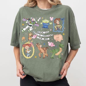 May include: An olive green t-shirt featuring a colorful graphic of characters from the animated movie Toy Story. The design includes Buzz Lightyear, Woody, and other characters, with the phrase "You've Got a Friend in Me."