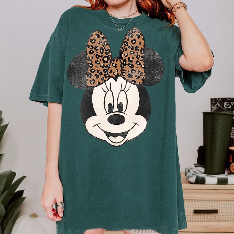 May include: Dark green t-shirt featuring a large Minnie Mouse graphic. The graphic includes a black and white face with a leopard print bow. The shirt has short sleeves and a relaxed fit.