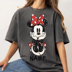 May include: Dark gray t-shirt featuring a Minnie Mouse graphic. The graphic shows Minnie Mouse in a seated pose, with a red bow with white polka dots. The word "NAME" is printed in black at the bottom of the graphic.