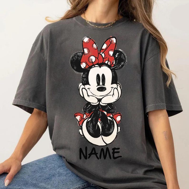 May include: Dark gray t-shirt featuring a Minnie Mouse graphic. The graphic shows Minnie Mouse in a seated pose, with a red bow with white polka dots. The word "NAME" is printed in black at the bottom of the graphic.