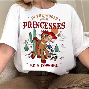 May include: White t-shirt with a graphic featuring a cartoon cowgirl hugging a horse, with a desert landscape backdrop. Text reads "In the world full of princesses, be a cowgirl."