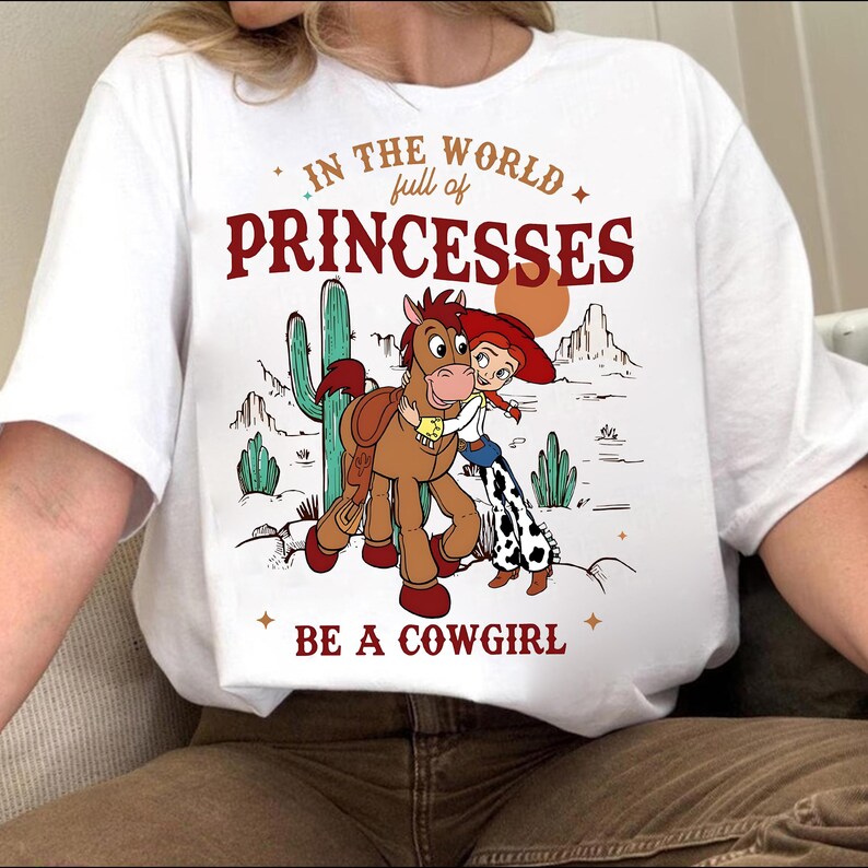May include: White t-shirt with a graphic featuring a cartoon cowgirl hugging a horse, with a desert landscape backdrop. Text reads "In the world full of princesses, be a cowgirl."
