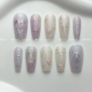 May include: A set of ten artificial nails in various shapes and designs. The nails feature a mix of pastel colors, including lavender, white, and pink. Embellishments include silver bows, hearts, stars, and celestial motifs. The brand name "Hani_nailbox" is visible.