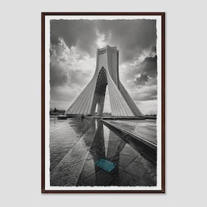 May include: A black and white framed print of the Azadi Tower in Tehran, featuring its distinct Y-shaped architectural structure. The monument is set against a dramatic cloudy sky, with a clear reflection visible on the wet stone ground below.