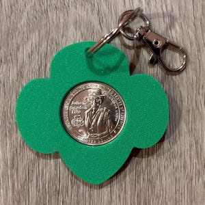 Può includere: Un portachiavi verde a forma di quadrifoglio con una moneta color argento raffigurante Juliette Gordon Low, fondatrice delle Girl Scouts. Il portachiavi ha un moschettone e un portachiavi argentati.