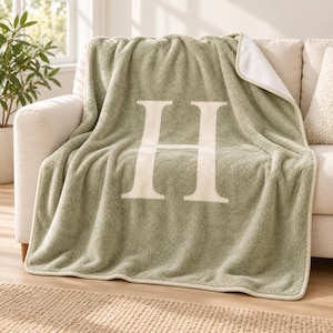 May include: A sage green throw blanket featuring a large, centered cream-colored letter H. The blanket has a soft, textured fleece-like appearance with a visible white underside folded over at the top corner and finished with a stitched edge.