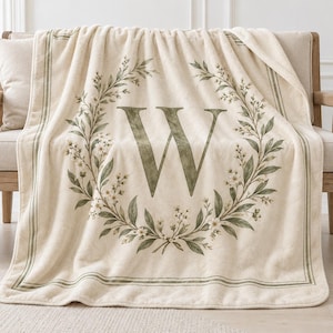 May include: A cream-colored plush throw blanket featuring a large, centered letter W in a dark green serif font. The letter is surrounded by a circular wreath of green leaves and small white flowers, framed by a thin green border along the edges.