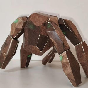 Puede incluir: Una escultura geométrica de papel marrón de una criatura con forma de cangrejo. La escultura tiene múltiples patas articuladas y un cuerpo segmentado. Se ven detalles verdes en las articulaciones. Los bordes están delineados en plata.