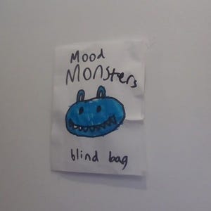 May include: A white paper bag with the words "Mood Monsters" and "blind bag" written in black marker. A blue monster with a smiling mouth is drawn on the bag.