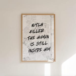 Op de afbeelding: Een ingelijste print met een licht houten frame. De achtergrond is een collage van krantenknipsels. Vetgedrukte zwarte tekst luidt "WITCH KILLER THE HUMAN IS STILL INSIDE HIM". De print is op een witte muur gemonteerd.